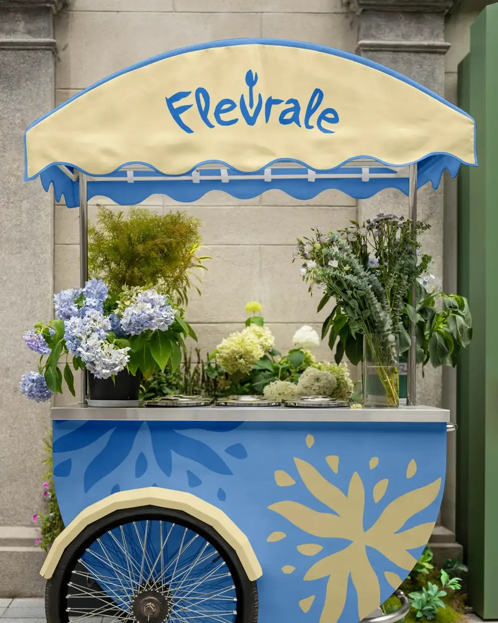 La charrette Fleurale, bleue et crème, chargée d'hortensias et d'eucalyptus devant un mur de pierre